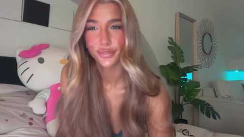 Video of a smiling, light-skinned woman with long, wavy blonde hair, wearing a pink shirt, sitting on a bed with a white Hello Kitty plush and a potted plant in the background.