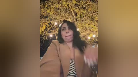 Video of a woman with shoulder-length dark hair, wearing a beige blazer over a striped top, standing outdoors near a lit-up tree, smiling.