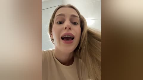 Video of a young, light-skinned woman with straight, shoulder-length blonde hair, wearing a beige T-shirt, smiling widely with her mouth open, in a bathroom with white tiled walls and a wooden door in the background.