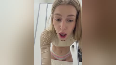 A video of a young, light-skinned woman with long blonde hair, wearing a cream long-sleeve top and colorful striped underwear, leaning over a mirror. She has a surprised expression.
