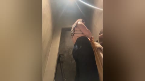 Video of a blonde woman in a black bra and pants, holding a light-colored garment, in a narrow, dimly lit hallway with a circular light overhead.