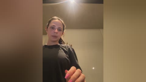 A video of a young woman with long brown hair, wearing a black top, taking a selfie in a dimly lit room with beige walls and a bright ceiling light.