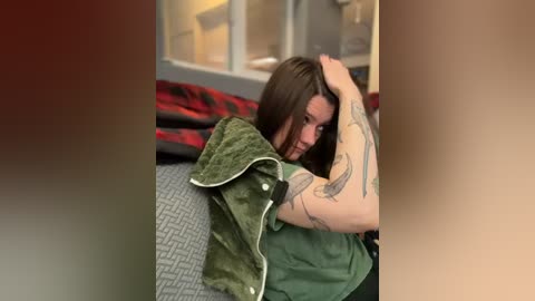A video of a woman with long brown hair, wearing a green shirt, lying on a bed with a green jacket and patterned blanket. She has tattoos on her arm. The background shows a window and beige walls.