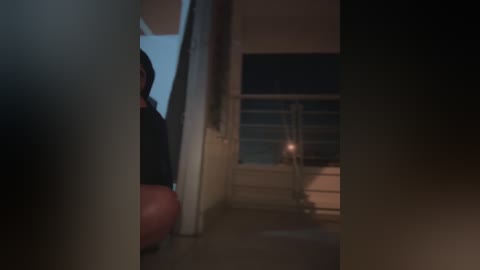 A dimly-lit, narrow hallway with a person's hand and shoulder visible in the foreground, leading to a partially open door with a faint light source inside.