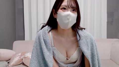 Video of an East Asian woman with long black hair, wearing a white mask, light pink bra, and grey cardigan, sitting on a pink couch with white curtains in the background.
