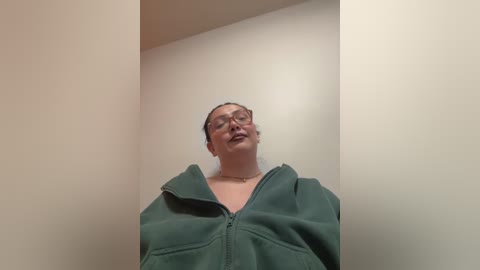 Video of a plus-sized, middle-aged woman with glasses and light skin, wearing a green jacket, standing in a beige-walled room with a slight downward angle.