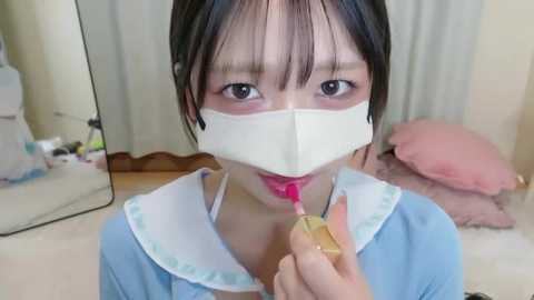 Video of an East Asian girl with light skin, dark hair, and bangs, wearing a light blue dress with a white collar. She has a white mask and is applying lipstick with a yellow tube. Background shows a pink pillow and a white bed.