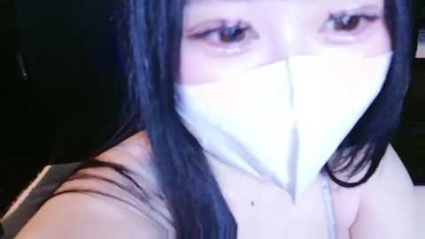 Media: Video of a woman with long black hair, wearing a white surgical mask, taken indoors with a dim background. She has a neutral expression and is dressed in a beige top.