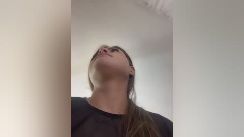 Video of a young woman with long, straight brown hair, wearing a black shirt, looking upward with her head tilted back, captured from a low angle, against a plain white background.