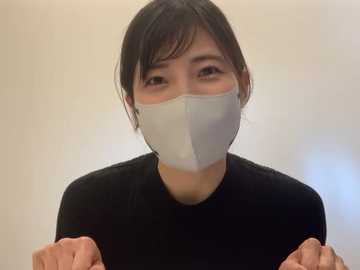 A video of an Asian woman with short black hair, wearing a white surgical mask, black top, and holding her hands up, indoors with a blurred, neutral background.