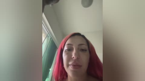 Video of a woman with fair skin and long red hair, wearing a red headscarf, looking directly at the camera in a dimly lit room.