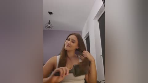 Video of a young woman with long brown hair, fair skin, and medium build, wearing a white tank top, using a hairdryer in a modern, minimalistic bathroom with white walls and a hanging light fixture.