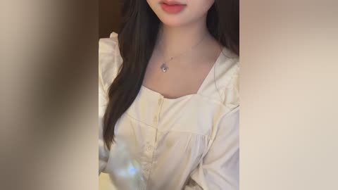 Video of a woman with fair skin, long dark hair, and wearing a white blouse with ruffled sleeves, smiling slightly. The background is blurred, featuring neutral tones.