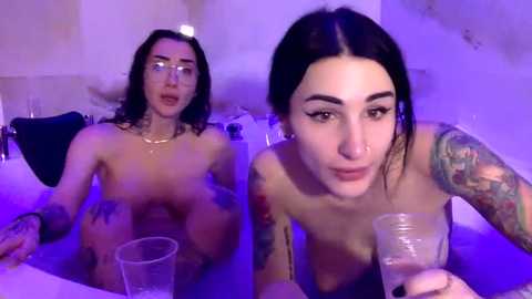 Video of two nude women in a bath, one with glasses, one with tattoos, both with fair skin, under purple lighting.