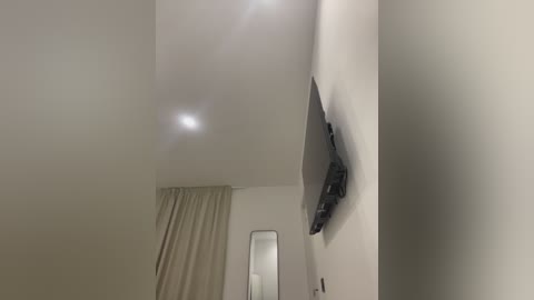 Video of a minimalist, modern bedroom with a flat-screen TV mounted on a white wall. The room features a beige curtain and a small mirror. The ceiling is white, and the overall decor is clean and simple.