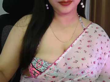 Video of a light-skinned woman with long black hair, wearing a sheer pink floral saree, exposing a colorful bra with geometric patterns, and dangling earrings.