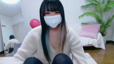 Video of an East Asian woman with long, black hair, wearing a white mask, a white cardigan, and black tights, kneeling on a white rug in a modern room with a pink balloon, a potted fern, and a bed in the background.