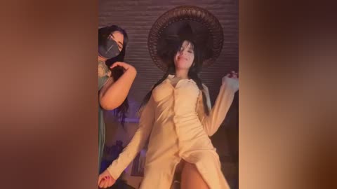 Video of two women in costumes: one with a black mask and green dress, the other in a beige jumpsuit with a large hat, dancing in a dimly lit room with a wooden ceiling.