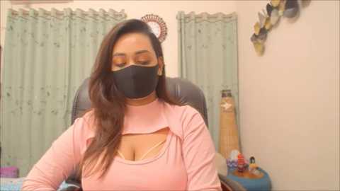 Video of a South Asian woman with medium skin tone, long brown hair, wearing a black face mask, pink long-sleeve top, and sitting in an office chair. Background shows green curtains, a desk with a bottle and a small plant, and a wall-mounted decorative item.