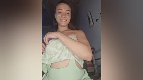 Video of a smiling, light-skinned woman with long brown hair, holding a light green shirt over her chest in a dimly lit room.