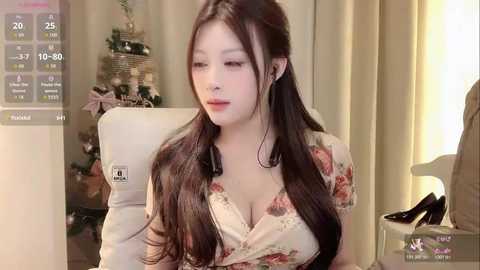 Video of a young Asian woman with long black hair, fair skin, wearing a low-cut floral blouse, seated in a white chair in a cozy living room.