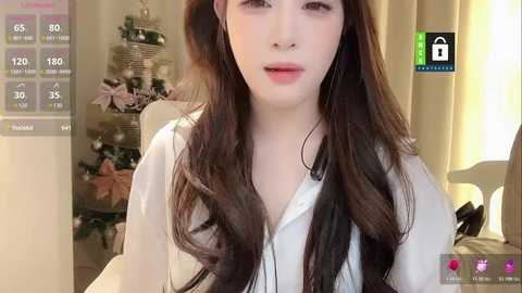 Video of a young Asian woman with long, wavy dark brown hair, wearing a white blouse, sitting in a room with Christmas decorations and a green screen in the background.