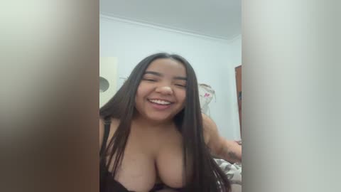 Media: A smiling, young woman with long dark hair, medium skin tone, and medium-sized breasts, wearing a black bra, seen from behind a vertical object in a softly lit room.