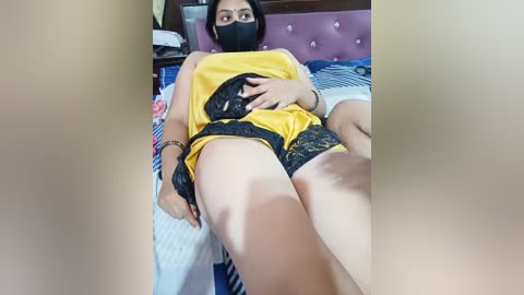 Video of a woman with medium skin tone, wearing a yellow top and black lace shorts, lying on a bed with a purple headboard, wearing a black face mask.