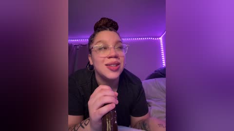 Video of a young, light-skinned woman with short, dark hair styled in a bun, wearing glasses and a black shirt, holding a beer bottle, in a dimly lit bedroom with purple LED strip lights.