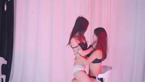 Video of two Asian women in matching black and white lingerie, embracing passionately in a studio setting with white curtains and red lighting.