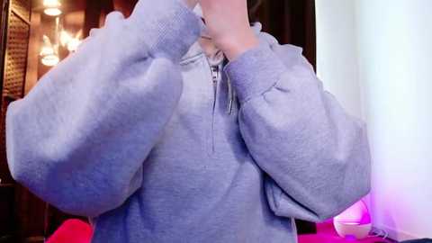 A video of a person in a grey hoodie, adjusting their collar, with a blurred background featuring a chandelier and a red couch.