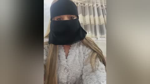 Video of a woman in a fluffy white bathrobe, wearing a black face mask with long blonde hair peeking out. She stands indoors with beige curtains in the background.