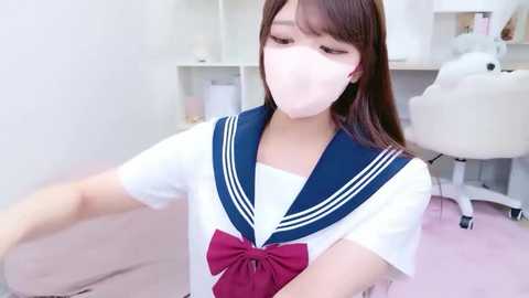Video of an East Asian woman with long brown hair, wearing a school uniform with a blue sailor collar and a red bow, standing indoors with a white chair and shelves in the background.