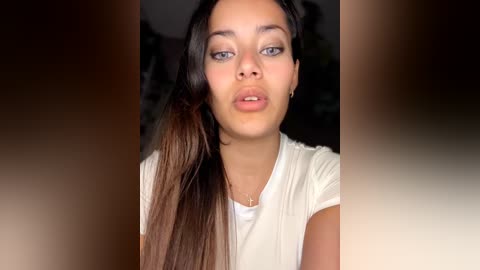 Video of a young woman with light skin and long dark hair, wearing a white top, making a kissy face, blurred background.