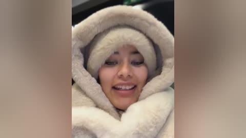 Video of a young, light-skinned Asian woman with a round face, wearing a cozy, cream-colored, fluffy blanket over her head, smiling. The background is blurred.