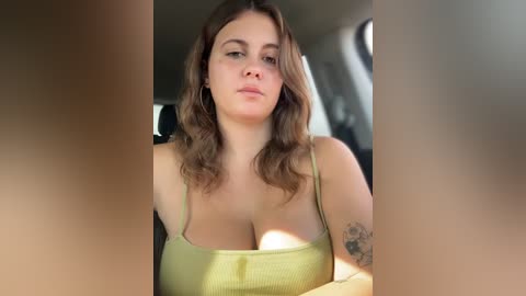 Video of a Caucasian woman with wavy brown hair, wearing a green tank top, sitting in a car. She has a tattoo on her right arm. The background shows a blurred interior.