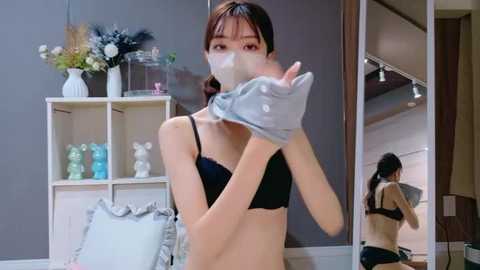 Video of an Asian woman in a black bra, removing a mask, in a modern living room with gray walls, white shelves, and a mirror.