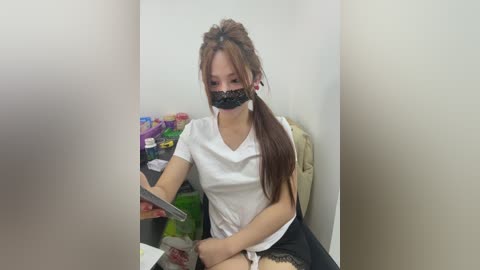 Video of a young Asian woman with long brown hair, wearing a white shirt, black lace mask, and black skirt, sitting at a table, holding a phone. Background shows a cluttered shelf with colorful items.