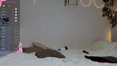 A video of a messy bedroom with white walls, a large plant, a pink plush toy, and two brown pillows on a white bed.