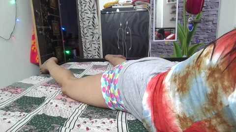 Media: Video of a woman lying face down on a patchwork quilted bed, wearing a tie-dye shirt and colorful patterned shorts, in a cozy, colorful room with a mix of modern and eclectic decor.