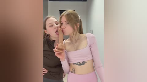 Video: Two young women, one licking a man's erect penis, wearing tight pink tops. Background: White hallway.