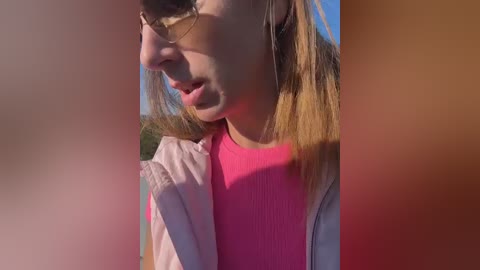 Video of a young woman with fair skin, shoulder-length blonde hair, wearing pink top and sunglasses, in a sunny outdoor setting.