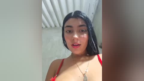 Video of a young woman with long, straight black hair, wearing red lingerie, standing in a bathroom with a white tiled wall and silver faucet.