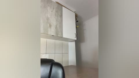 Video of a minimalist kitchen in progress, featuring white cabinets with a concrete backsplash, light beige walls, and a black leather chair partially visible on the left.