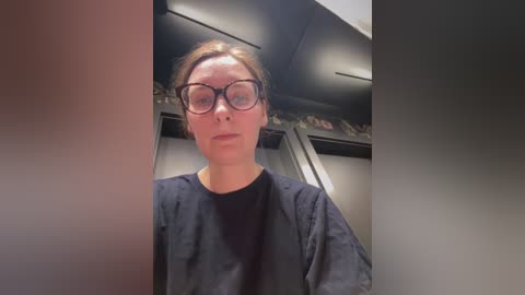 Video of a middle-aged woman with glasses and light skin, wearing a black robe, standing in a dimly lit, modern room with a wall of cabinets and a shelf displaying shoes in the background.