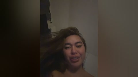 Video of a young woman with long brown hair, fair skin, and dark eyebrows, laughing while looking into a mirror. The background is dimly lit, suggesting an indoor setting with minimal decor.