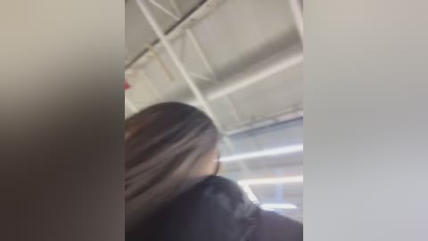 A blurry video of a person with long, straight, light brown hair, facing away, in a dimly lit, industrial setting with white ceiling beams and fluorescent lighting.