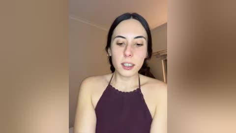 Video of a young woman with fair skin and dark hair in a braid, wearing a dark halter top, indoors with beige walls and a window.