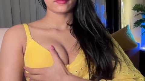 Video of a woman with long black hair, wearing a yellow lace bra, covering her cleavage with her hand. She has light skin and is indoors, next to a plant and a cushion.