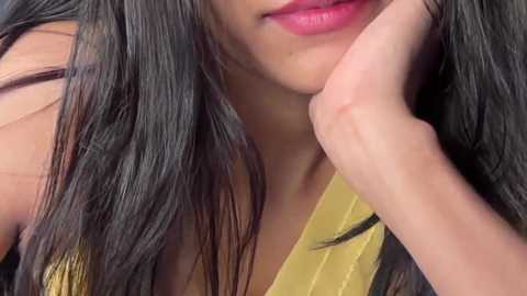 Video of a young woman with long, straight black hair, light brown skin, and full, pink lips. She's wearing a yellow top and resting her chin on her hand.